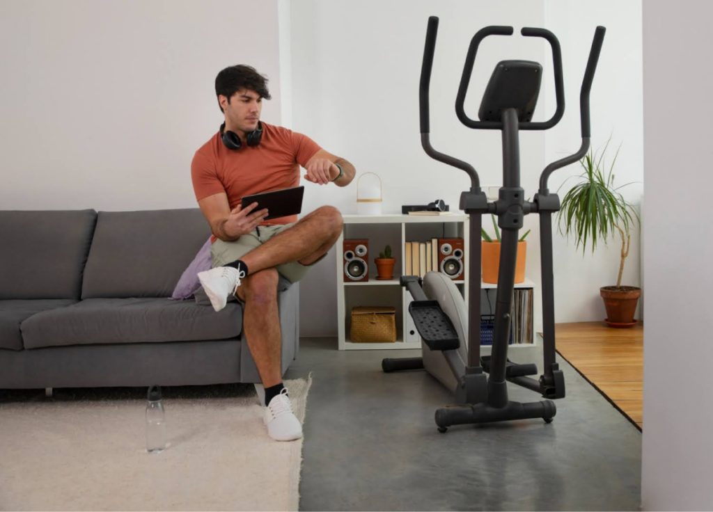 A man checks his watch while sitting on a couch with an elliptical nearby, showcasing fitness integration in home spaces.