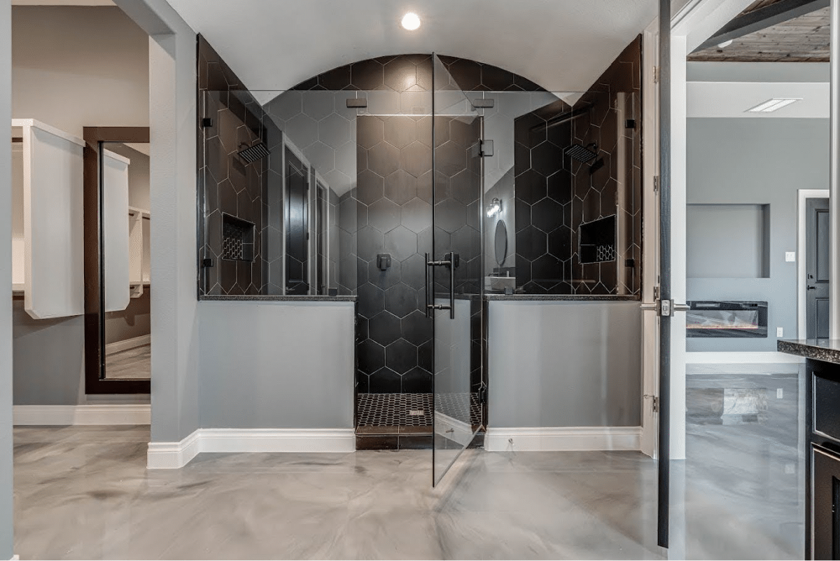 A modern bathroom with a large glass shower enclosure, black hexagonal tiles, and a metallic floor.