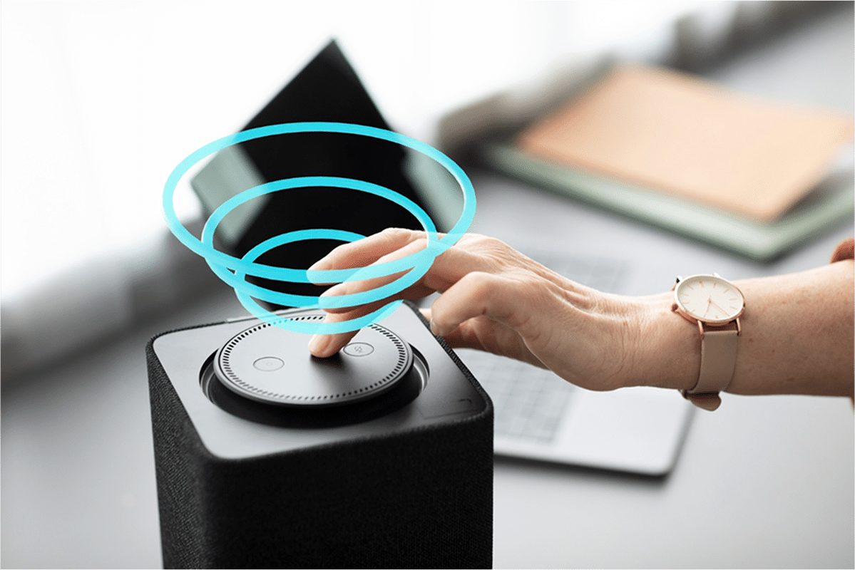 Hand pressing button on black smart speaker, activating it.