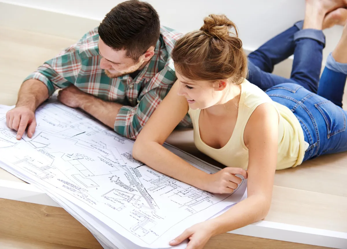 A man and woman lying on the floor, studying a large house blueprint together.