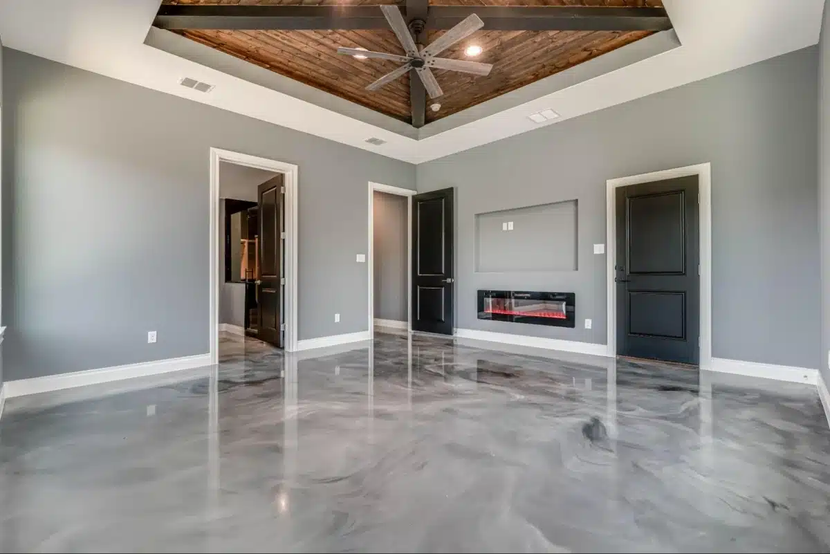 A spacious bedroom featuring a wooden-inset tray ceiling with a large fan, gray walls, black doors, a built-in electric fireplace, and a polished epoxy floor.