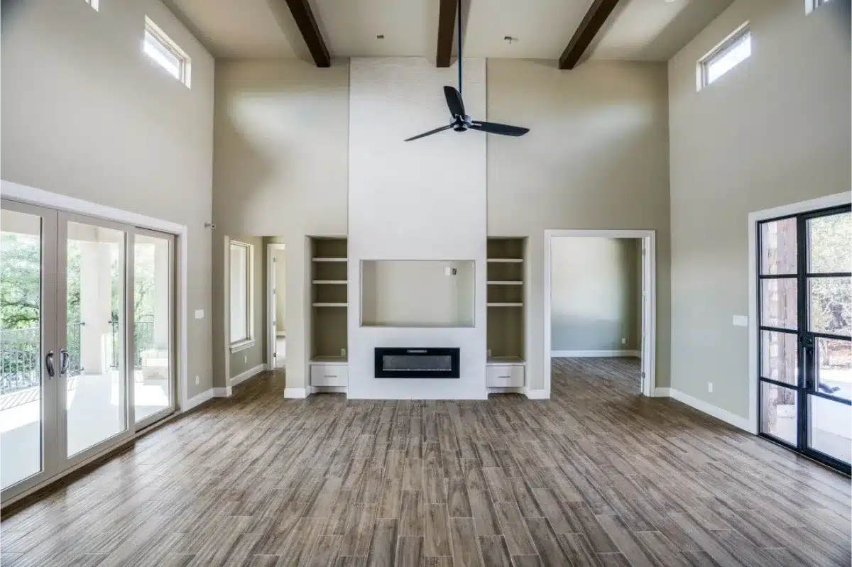 An open living room featuring a floor-to-ceiling fireplace, built-in shelving, high ceilings with exposed beams, and wood-look tile flooring with ample natural light.