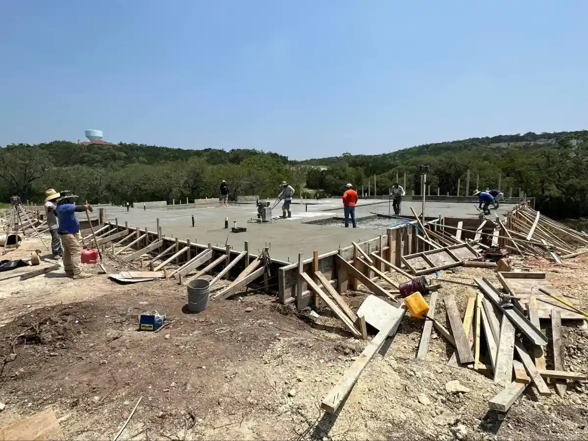 Construction workers smoothing freshly poured concrete on a house foundation in a rural setting.