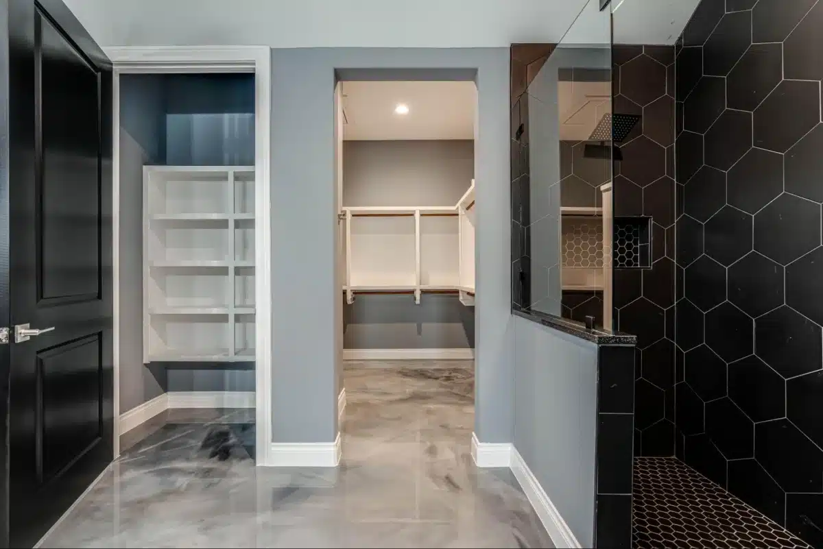 A bathroom with a black hexagonal-tiled shower, gray walls, an epoxy floor, and an open passage leading to a walk-in closet with built-in shelving.